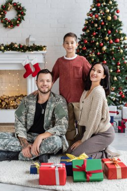Karısı ve oğlu olan mutlu bir asker hediye kutularının ve süslü Noel ağacının yanındaki kameraya bakıyor.