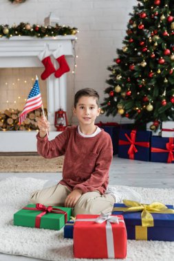 Amerika bayrağıyla gülümseyen çocuk oturma odasında hediye kutularının yanında oturmuş Noel ağacı ve süslü şömineli.