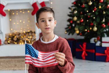 Küçük Amerikan bayrağıyla vatansever çocuk bulanık oturma odasında Noel dekoruyla kameraya bakıyor.