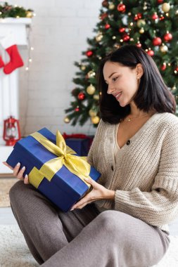 Mutlu esmer kadın bulanık noel ağacının yanında sarı kurdeleli mavi hediye kutusuna bakıyor.