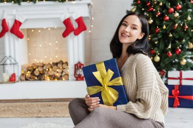 Noel ağacının yanında hediye kutusuyla oturan neşeli bir kadın ve evinde süslü bir şömine.