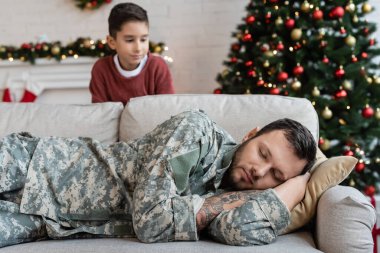 Bulanık çocuk kamuflajlı bitkin babasına bakıyor oturma odasındaki kanepede uyuyor ve Noel süslemeleri yapıyor.