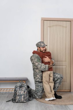 Mutlu bir asker oğlunu sırt çantası ve eve giriş kapısının yanında kucaklıyor.