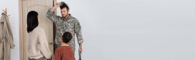 man in camouflage taking off military cap near wife and son meeting him at home, banner clipart
