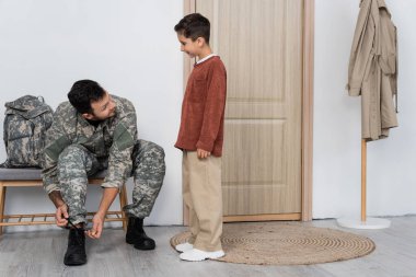 gülümseyen çocuk evde sırt çantasının yanında otururken asker üniforması içinde babasına bakıyor.