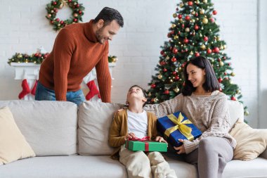 Mutlu çocuk, Noel hediyeleri ve gülümseyen anneyle koltukta oturmuş babama bakıyor.