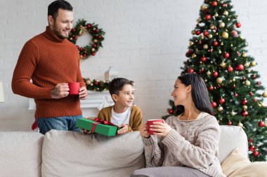 Mutlu çocuk, ailesinin yanında elinde sıcak kakao bardaklarıyla Noel hediyesi tutuyor.