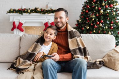 Neşeli baba ve oğul sıcak battaniyenin altında kanepede oturuyor ve dekore edilmiş Noel ağacının yanında televizyon seyrediyorlar.