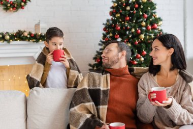 Çocuk Noel ağacının yanında sıcak kakao içiyor ve ebeveynler sıcak battaniyenin altında koltukta oturuyor.