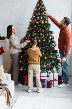 Aile, oturma odasındaki hediye kutularının yanında noel balolarıyla ladin süslemesi yapıyor.