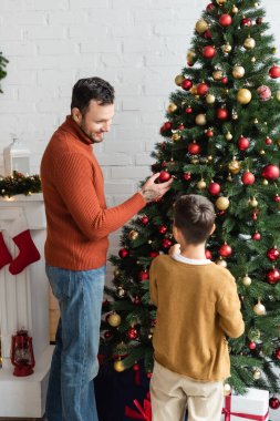 Gülümseyen adam, evdeki süslemelerle Noel ağacını süslerken oğluna bakıyor.