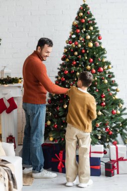 Mutlu baba ve oğlu yeşil çam ağacını hediye kutularının yanında noel toplarıyla süslüyorlar.