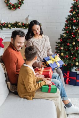 Mutlu bir aile, hediye kutularıyla evdeki koltukta oturmuş Noel ağacının yanında.