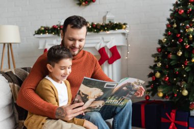 Gülümseyen baba ve oğul oturma odasında Noel dekoru ile dergi okuyorlar.