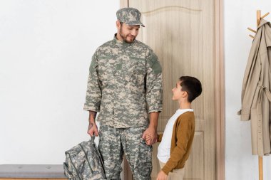 boy and father in camouflage holding hands and smiling at each other at home clipart