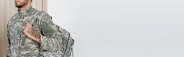 cropped view of military man with beard and tattoo holding backpack at home, banner clipart