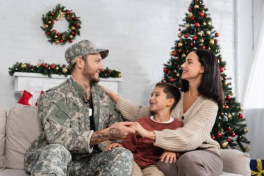 Mutlu kadın kocasının elini tutuyor. Askeri üniformalı. Mutlu oğul ve noel ağacının yanında.