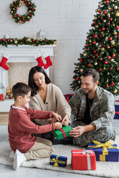 мужчина в камуфляже готовит подарки с женой и сыном возле рождественской елки