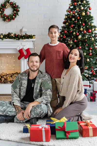 happy military man with wife and son looking at camera near gift boxes and decorated christmas tree