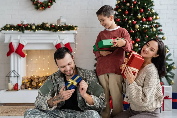 smiling military man with son and wife holding gift boxes near christmas tree and decorated fireplace