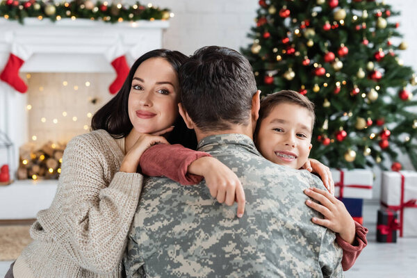 счастливая мать и сын обнимают мужчину в камуфляже возле размытой рождественской елки и украшенного камина