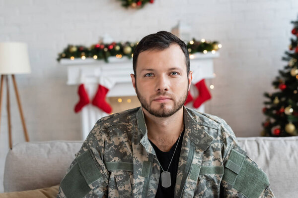 serious unshaven military man looking at camera near fireplace with christmas decor on blurred background