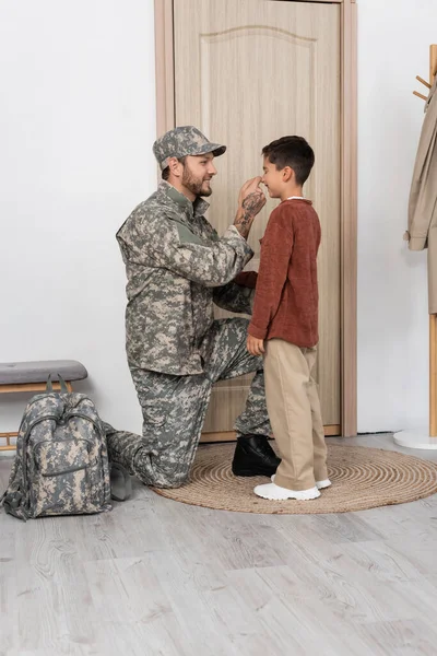 happy man in camouflage touching nose of son meeting him near door at home