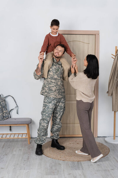 smiling boy piggybacking dad in military uniform near wife and entrance door at home