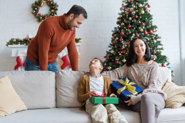 幸せな男は息子を見てソファの上に座ってお母さんとクリスマスプレゼント — ストック写真