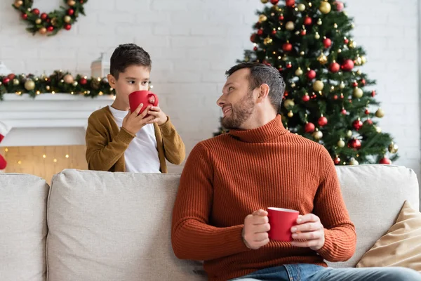 Cheerful Man Sitting Couch Looking Son Drinking Warm Cocoa Decorated — Stock Photo, Image