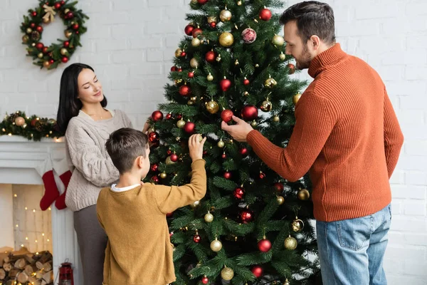 happy family decorating green spruce with christmas balls in living room with fireplace