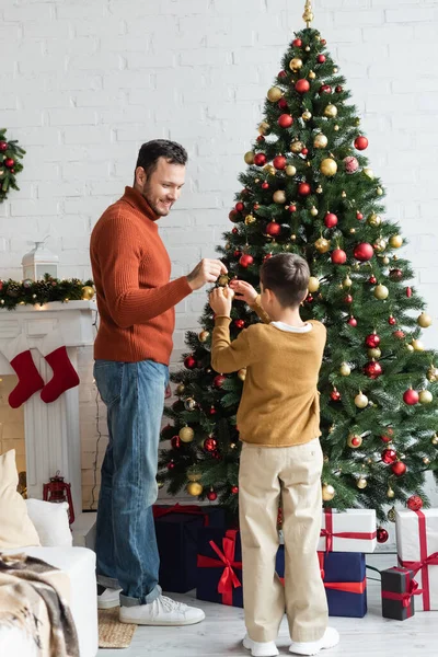 Mutlu adam oturma odasındaki hediye kutularının yanında oğluyla Noel ağacı süslüyor.