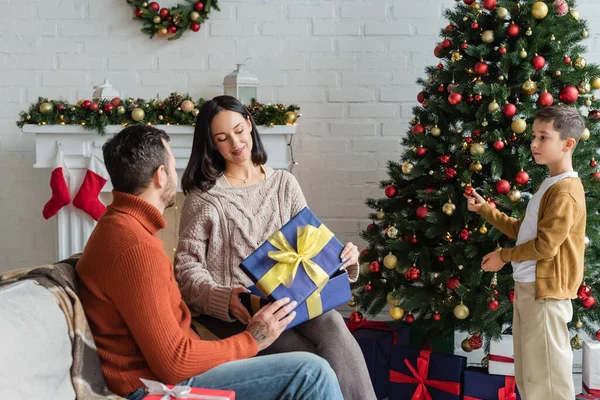 夫と息子の近くにギフトボックスを開く笑顔クリスマスツリーを飾る妻 — ストック写真