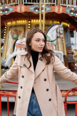 Bej renkli trençkotlu güzel bir kadın lunaparkta atlı karıncaların yanında duruyor.