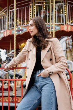 Bej renkli trençkotlu ve kot pantolonlu genç bir kadın lunaparkta atlıkarıncada el ele duruyor.