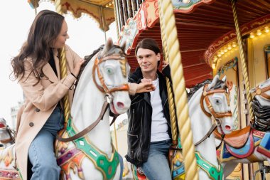 cheerful young couple in stylish outfits holding hands and riding carousel horses in amusement park clipart