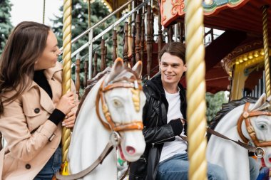 Neşeli genç adam eğlence parkında atlı karıncaya binen kız arkadaşına bakıyor.