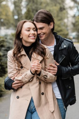 joyful young man hugging happy girlfriend in trench coat outdoors clipart
