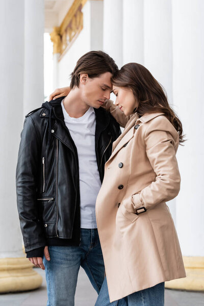 pretty young woman with hand in pocket of trench coat hugging boyfriend in stylish jacket near white columns