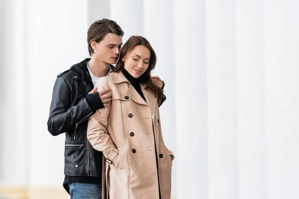 时尚男人 穿着夹克 抱着快乐的女朋友的肩膀 手插在雪白的柱子旁的战壕外套口袋里 — 图库照片