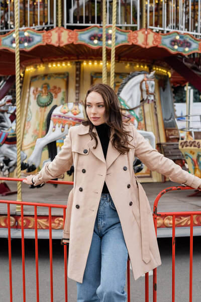 pretty young woman in beige trench coat and jeans standing near carousel in amusement park