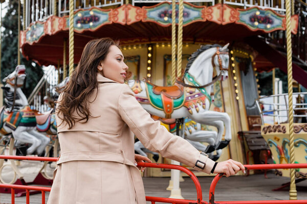 side view of curly young woman in beige trench coat standing near carousel in amusement park