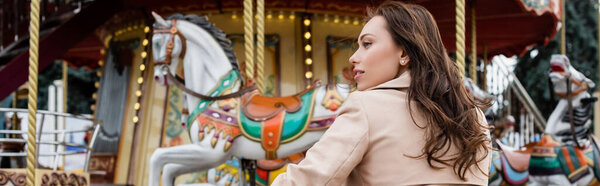 side view of curly young woman in beige trench coat standing near carousel in amusement park, banner