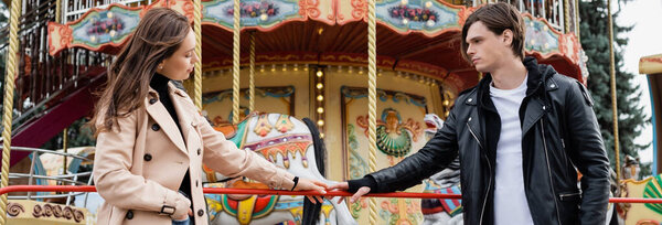 young couple standing near carousel and touching hands in amusement park, banner