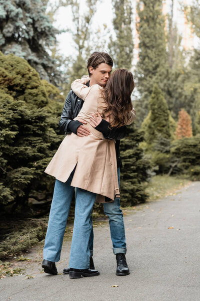 full length of young man dancing with brunette girlfriend in trench coat in autumnal park 