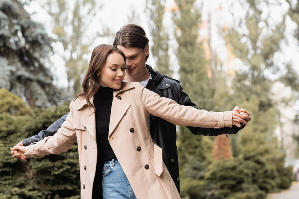 cheerful man holding hands with pleased girlfriend in beige trench coat 