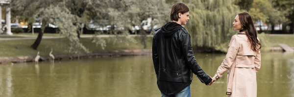 молодая пара в осенних нарядах, держась за руки, стоя возле озера в парке, баннер