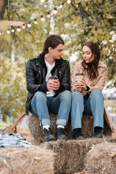 young woman and man in stylish outfits holding paper cups and sitting in autumnal park