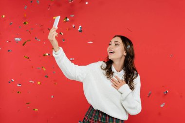 Süveter giyen neşeli kadın kırmızı arka planda konfeti altında selfie çekiyor. 