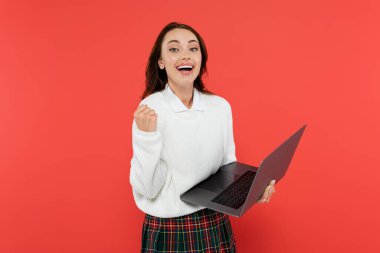 Excited woman in warm jumper showing yes gesture and holding laptop isolated on red  clipart